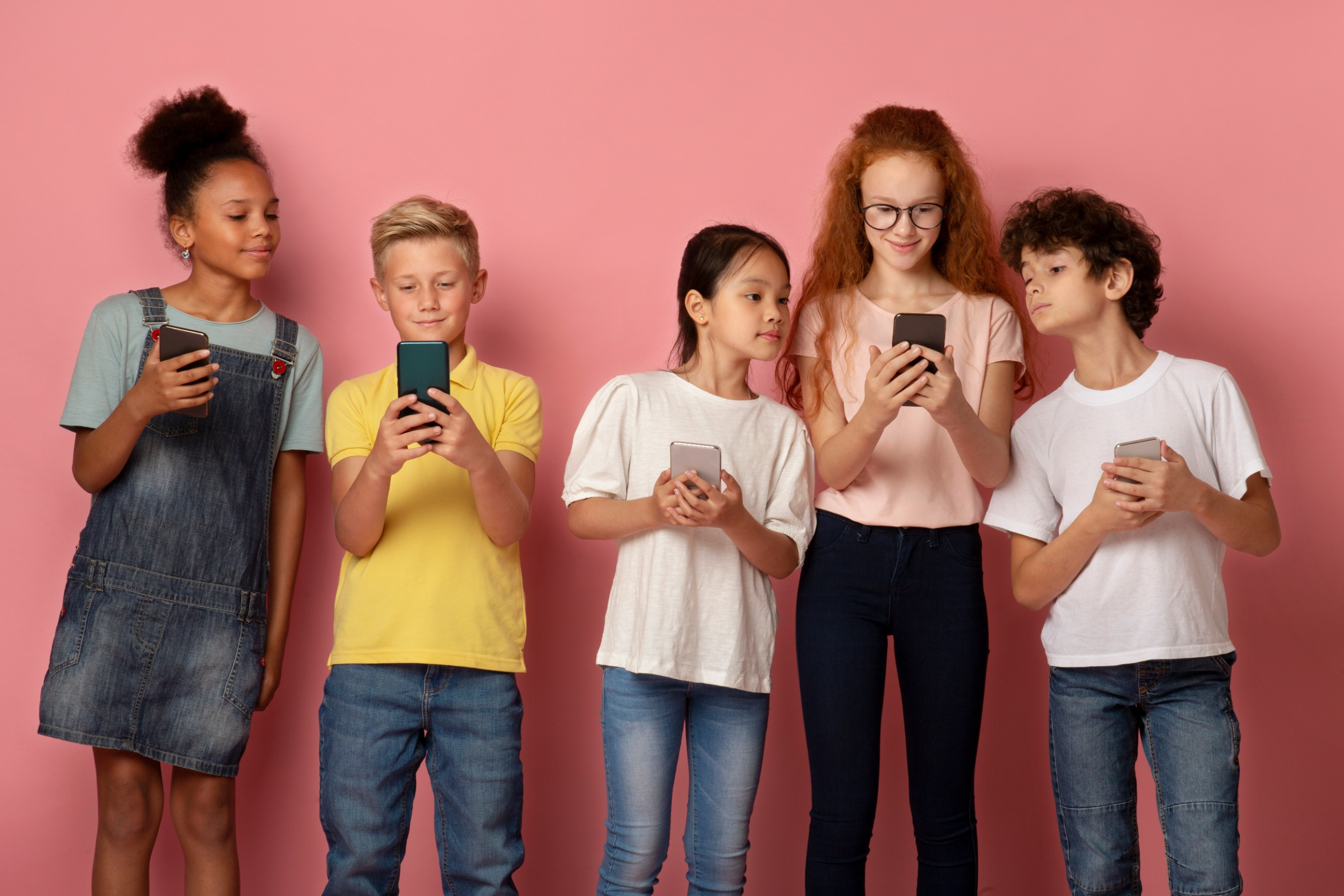 Group of children using mobile phones, looking at screen and smiling, standing against pink background