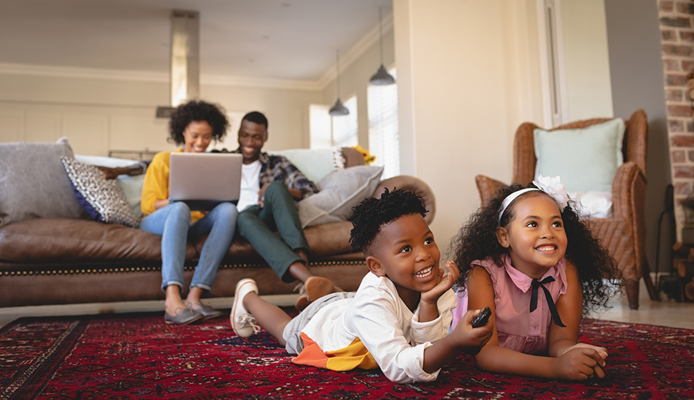 African American sibling lying on floor and watching television while parents using laptop on sofa in a comfortable home