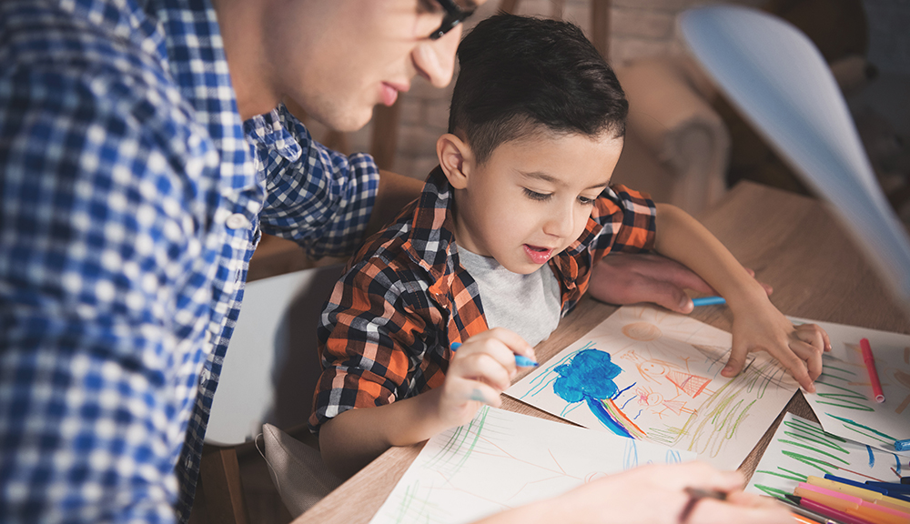 Father is helping son to draw with color pencils and markers on paper at table at night at home.