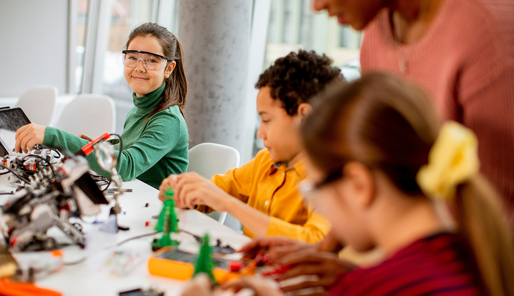 Smiling African American female science teacher with group of kids programming electric toys and robots at robotics classroom
